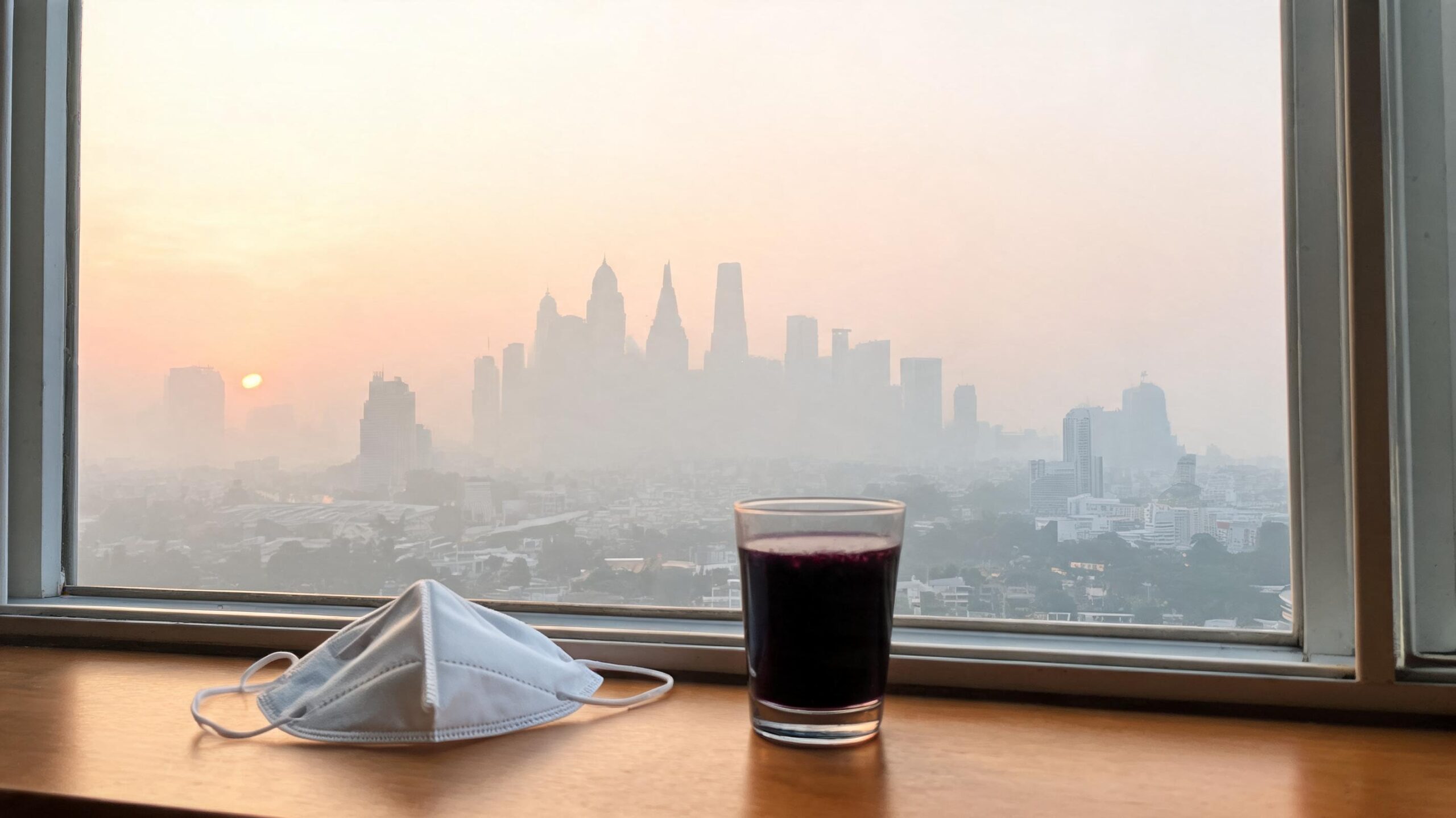 Hazy urban skyline at dawn seen through a window with an N95 mask and a glass of deep purple-black berry juice on a wooden windowsill.
