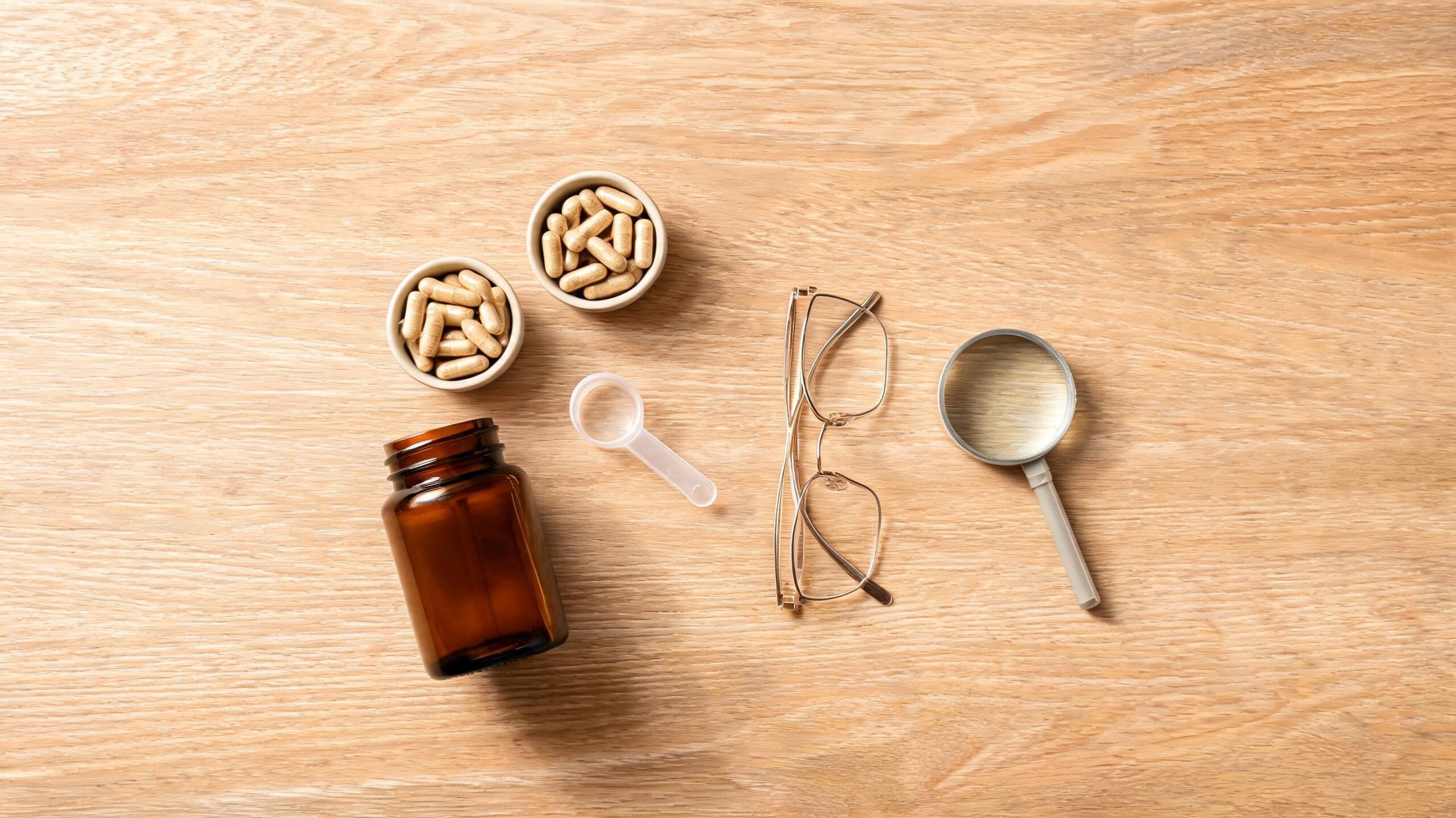 Flat lay of joint supplement capsules and a powder scoop on a light wooden table with reading glasses